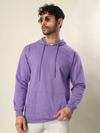 A man wearing a lavender purple pullover hoodie with matching drawstrings, paired with white pants, standing against a plain light beige background.