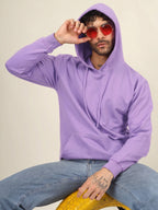 A man wearing a lavender purple hoodie and light blue jeans, sitting on a yellow textured stool against a plain beige background. He is wearing round red-tinted sunglasses and holding one up to his eye.