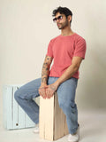 Man wearing a pink t-shirt and blue jeans sitting on a wooden box against a plain background