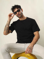 Man wearing a black t-shirt and sunglasses, sitting on a yellow object against a neutral background