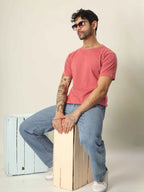 Man wearing a pink t-shirt and blue jeans sitting on a wooden box against a plain background