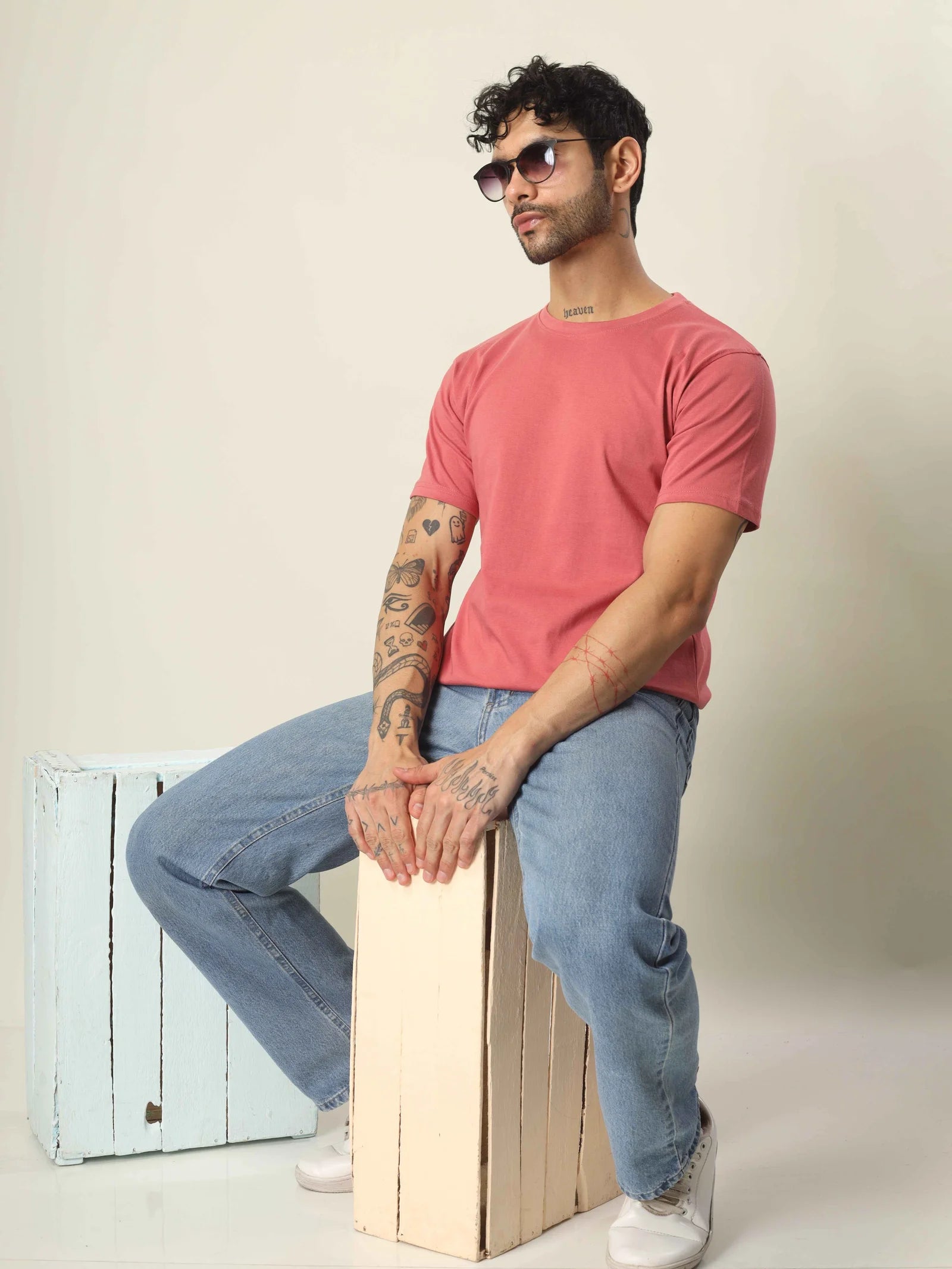 Man wearing a pink t-shirt and blue jeans sitting on a wooden box against a plain background