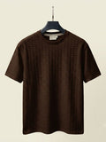 A dark brown short-sleeve crewneck t-shirt with a textured waffle pattern, hanging on a black hanger against a light beige background.