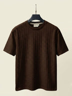 A dark brown short-sleeve crewneck t-shirt with a textured waffle pattern, hanging on a black hanger against a light beige background.