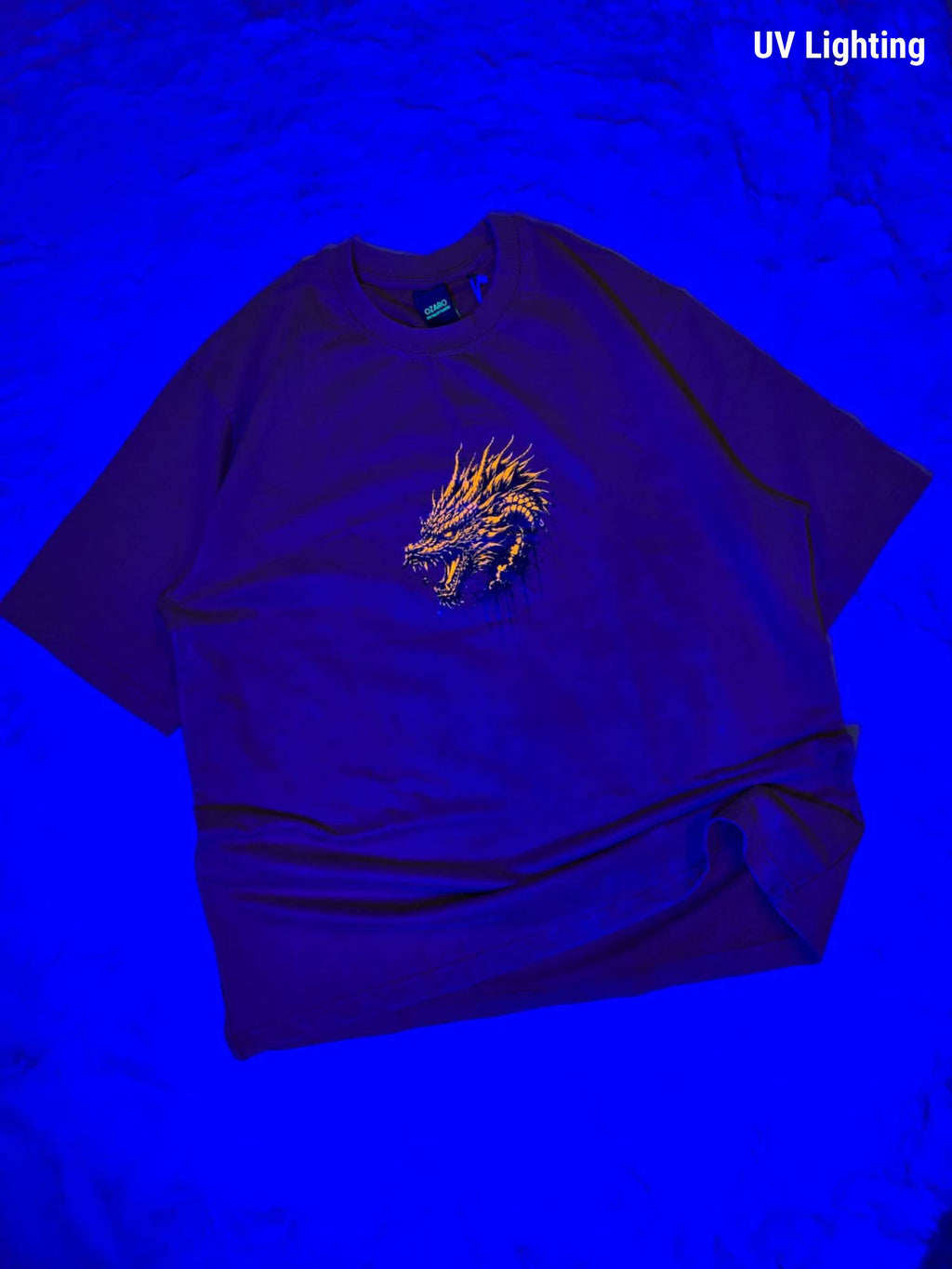 A dark purple t-shirt with a glowing yellow-orange dragon graphic on the chest, displayed under blue UV lighting on a textured blue background. The text 'UV Lighting' is visible in the top right corner.