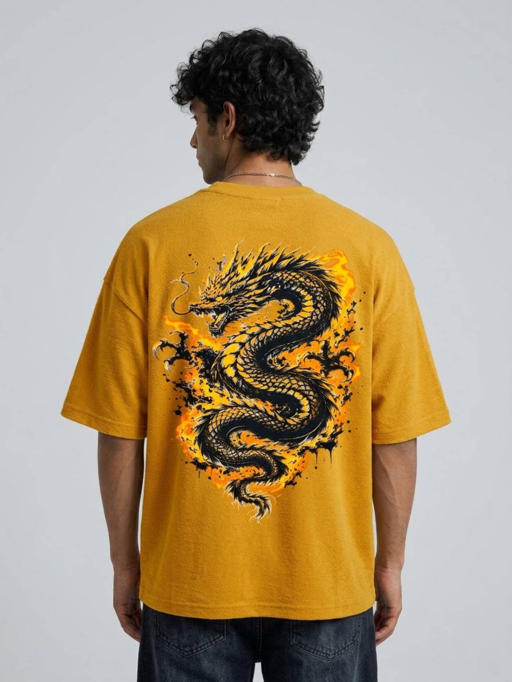 A man viewed from behind wearing a mustard yellow t-shirt with a large black and orange dragon graphic printed on the back, paired with dark blue jeans against a light gray studio background.