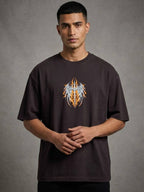 A man wearing a dark brown t-shirt with a graphic of a stylized winged skull in orange, yellow, and white, standing against a textured gray background.