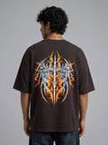A man viewed from behind wearing a dark brown oversized t-shirt with a large graphic print on the back. The graphic features a silver winged sword design surrounded by orange and yellow flames.