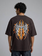 A man viewed from behind wearing a dark brown oversized t-shirt with a large graphic print on the back. The graphic features a silver winged sword design surrounded by orange and yellow flames.